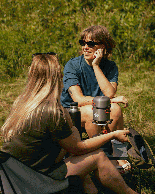 Two friends camping beside a lake with a black tent, folding chairs and a portable stove, enjoying drinks outdoors