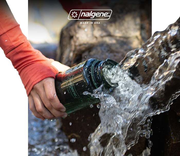 Hand holding green Nalgene bottle being filled from a mountain stream beside rocks, Nalgene logo visible
