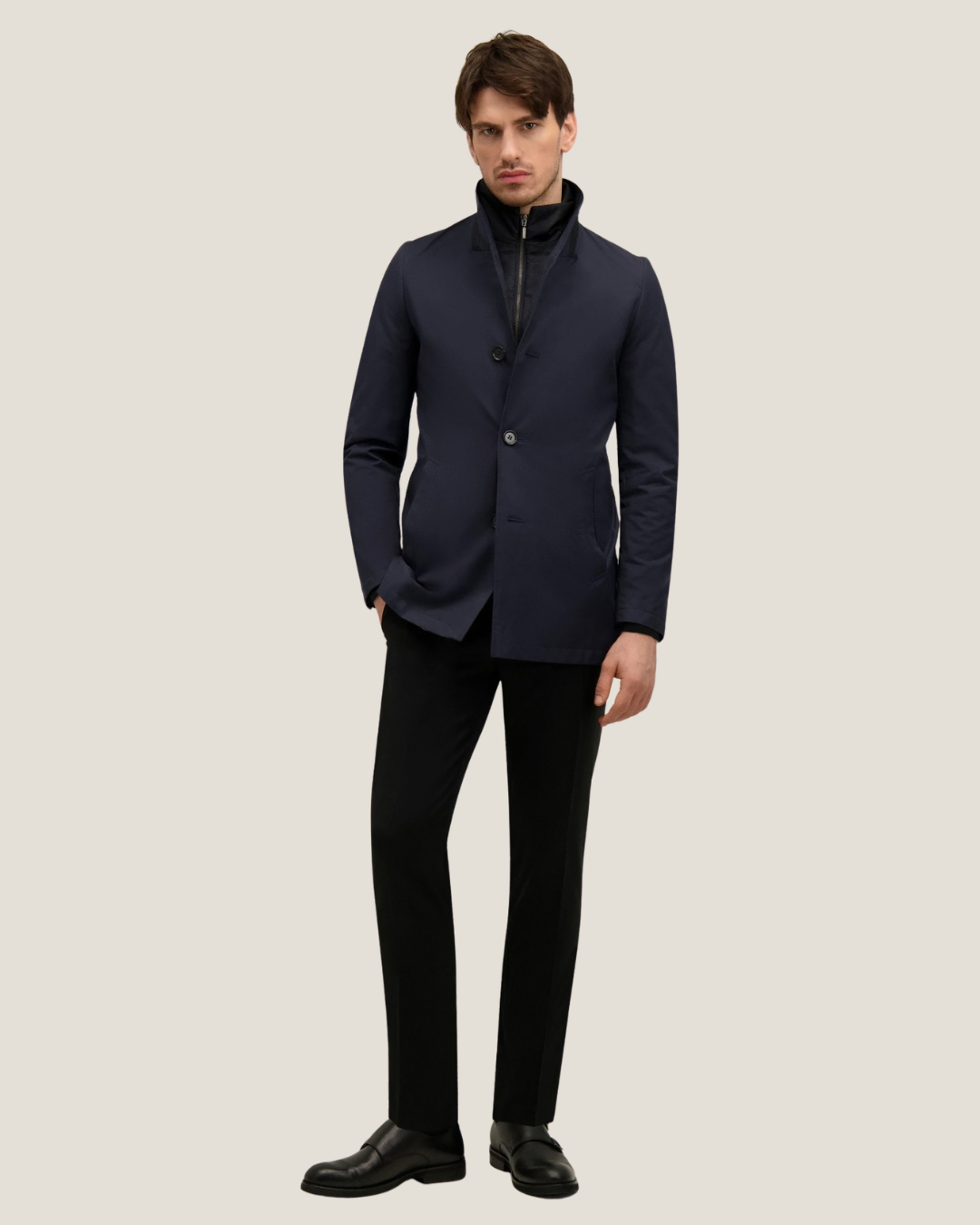 Male model wearing navy tailored jacket over a black zip-up layer, black trousers and leather loafers on a neutral background