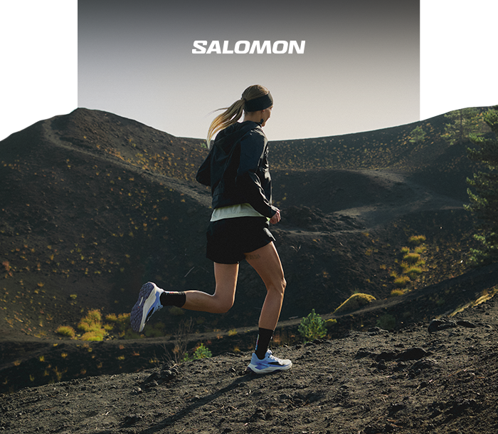 Woman trail running on volcanic terrain wearing Salomon shoes and jacket, running past rugged black hills under a grey sky
