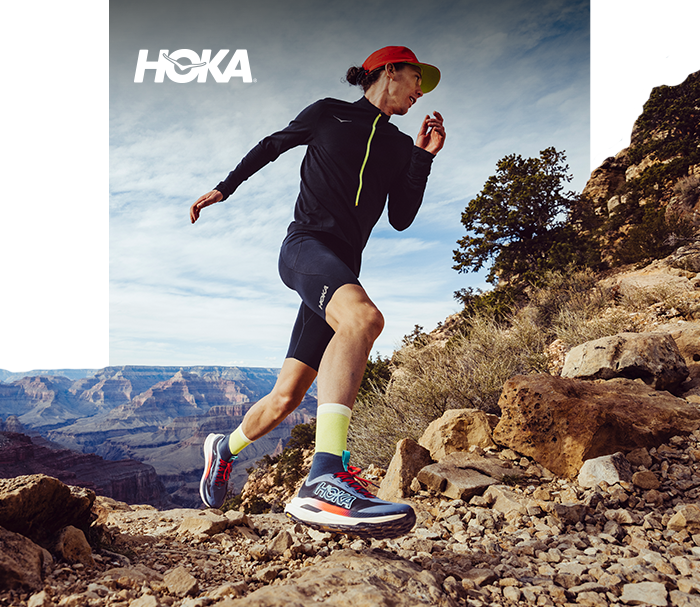 Trail runner wearing HOKA shoes and bright cap running on a rocky canyon path under a dramatic sky