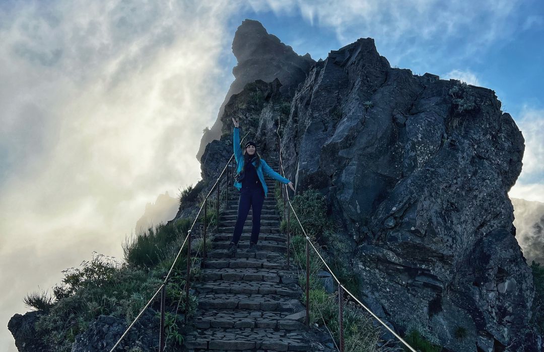 Hiker in blue jacket raising an arm on a steep stone mountain staircase beside a jagged rock face with misty clouds