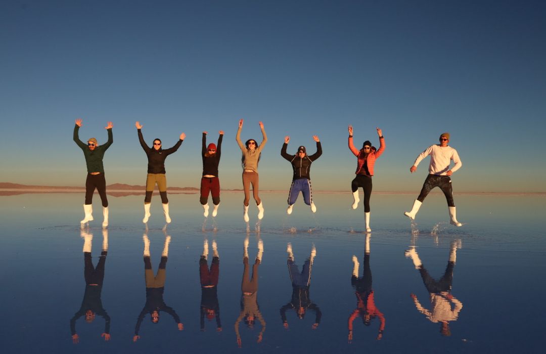 Seven people jumping on a mirror-like salt flat at sunset with clear reflections and colourful clothing