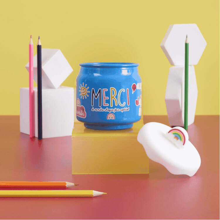 Patterned grid of colourful stationery still life with "MERCI" jars, pencils, geometric blocks and rainbow cloud erasers on a yellow backdrop