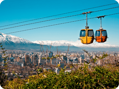 Two colourful cable cars above a city skyline with snow‑capped mountains in the background, viewed from a green hillside