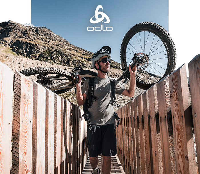 Helmeted man carrying a mountain bike across a wooden bridge with rocky alpine backdrop, wearing Odlo sportswear