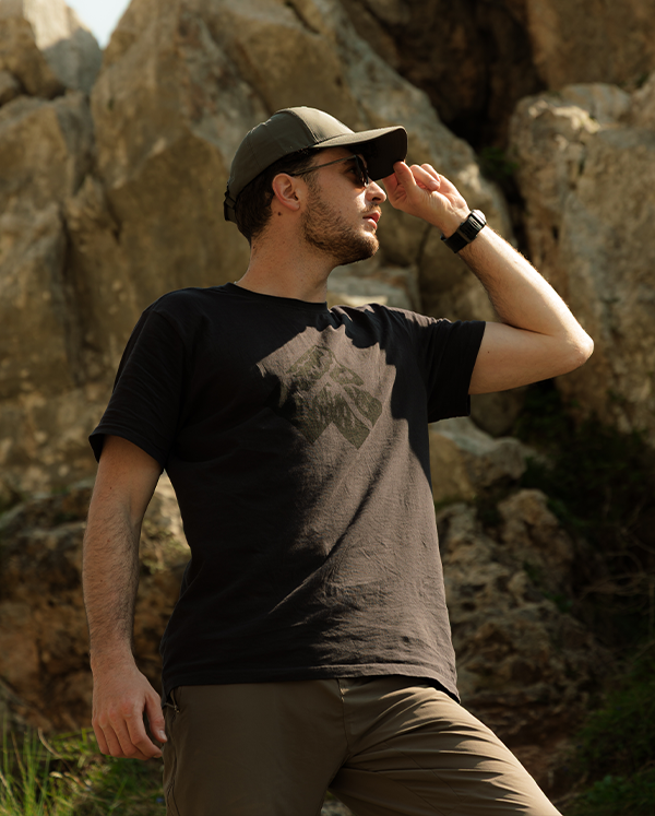 Young man in cap and sunglasses adjusting his cap on a rocky hillside, wearing a black t-shirt and outdoor trousers