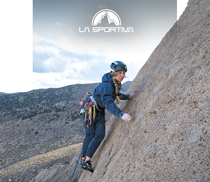 Climber in helmet and harness ascending a steep granite slab, La Sportiva logo overhead, rugged mountain landscape
