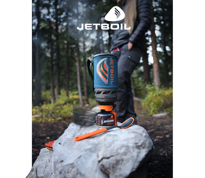 Jetboil camping stove on a rock with orange spork and fuel canister at a pine forest campsite, blurred hiker in background