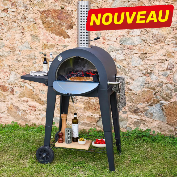 Portable wood-fired outdoor pizza oven on wheeled stand with pizza baking, set on grass against a stone wall