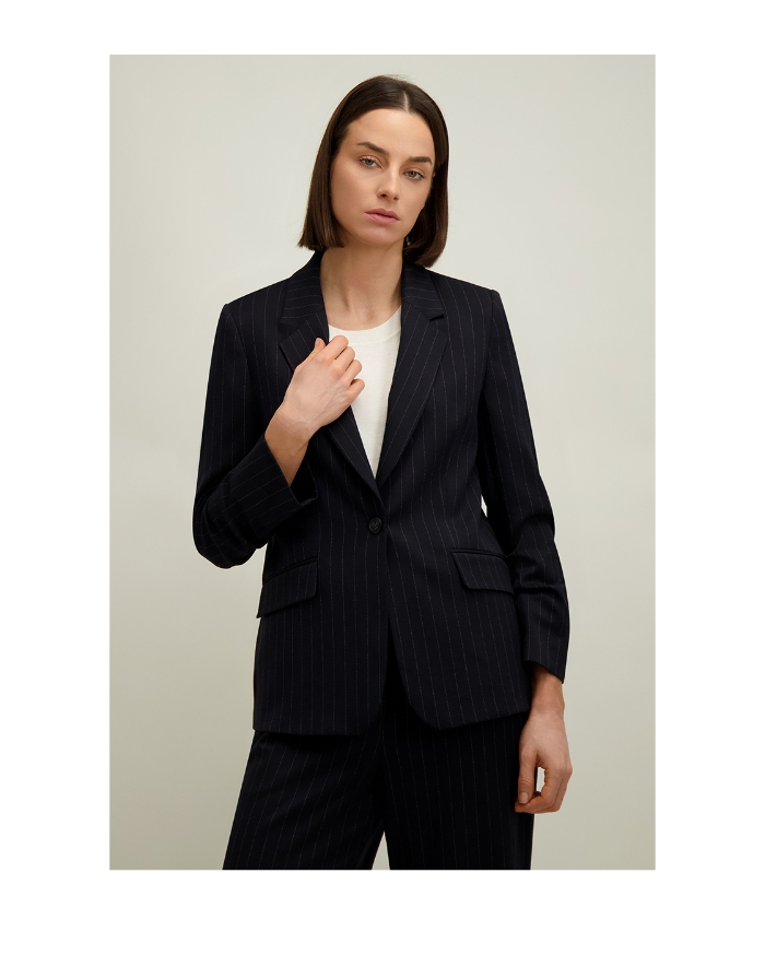 Model with bob haircut wearing navy pinstripe blazer and matching trousers over white T‑shirt, hand on lapel against neutral background