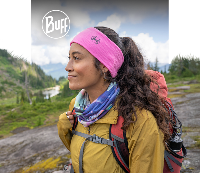 Hiker wearing pink Buff headband, colourful neck gaiter, yellow jacket and red backpack on mountain trail under cloudy sky