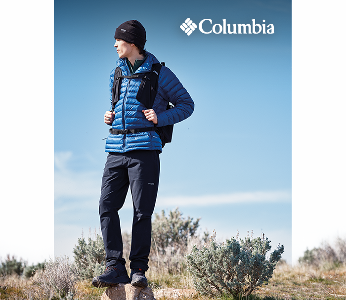 Hiker in blue Columbia puffer jacket and backpack standing on a rock overlooking scrubland under clear blue sky, Columbia logo