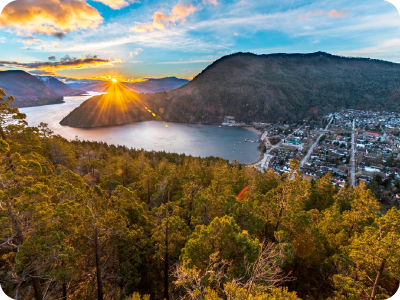 Sunrise sunburst over a mountain lake, dense forest in the foreground and a waterfront town along the shoreline under a colourful sky