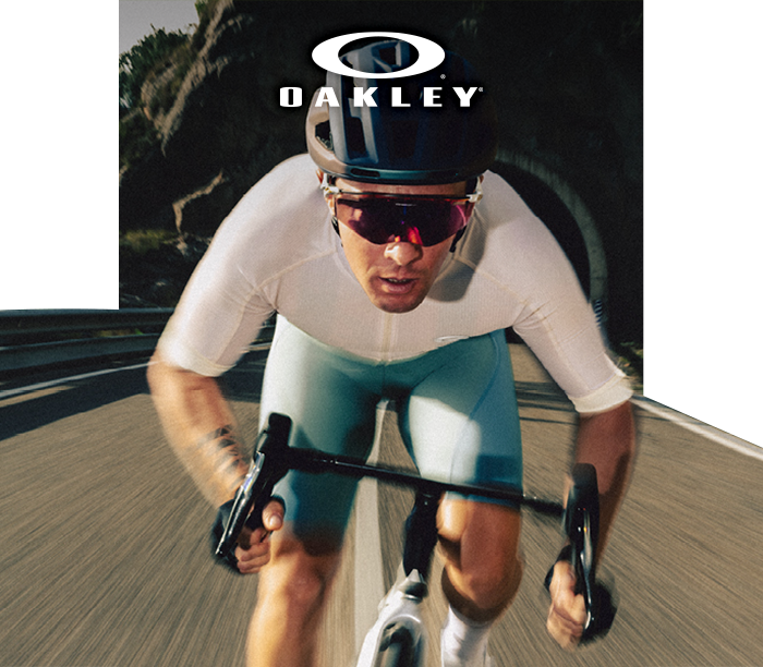 Cyclist wearing Oakley helmet and mirrored sunglasses racing on a road bike down a winding road with Oakley logo above