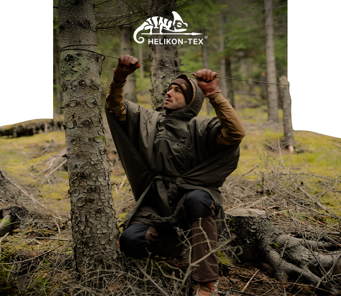 Man in olive Helikon‑Tex poncho securing a guyline between trees in a mossy conifer forest, camping shelter setup