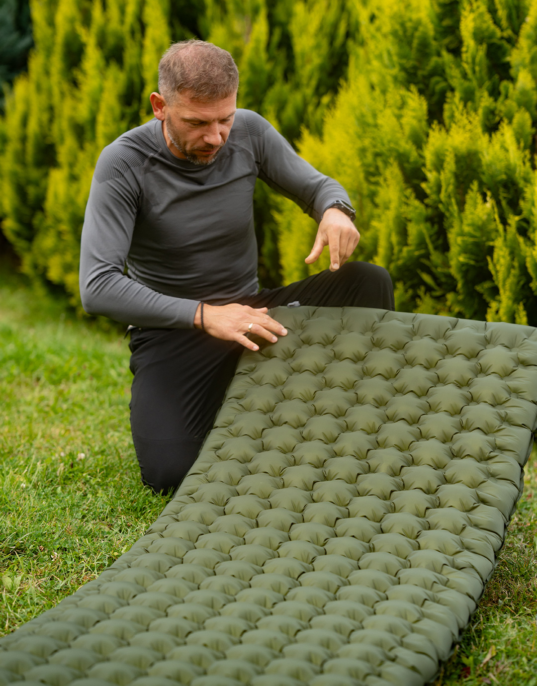 Man kneeling on grass inspects green inflatable camping mattress with tufted air cells in a garden