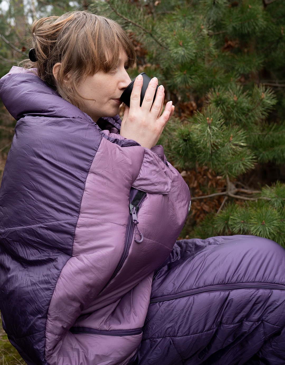 Person wrapped in a purple sleeping-bag jacket drinking from a mug while seated outdoors beside pine trees