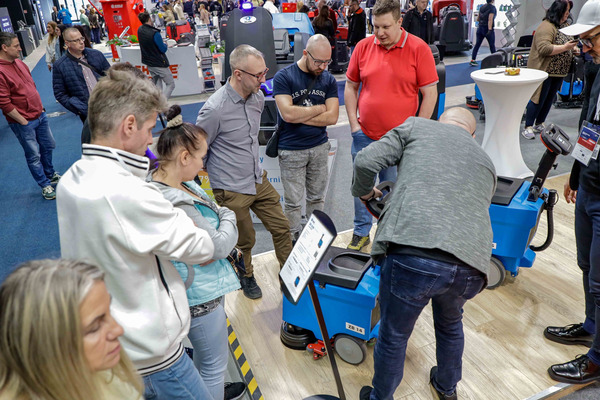 Visitors gathered around a demonstrator showing a blue industrial floor‑cleaning machine at a busy trade show booth