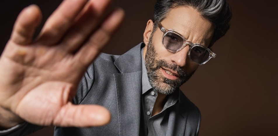 Close-up of a bearded man in a grey suit and clear-framed glasses reaching his hand towards the camera against a brown background