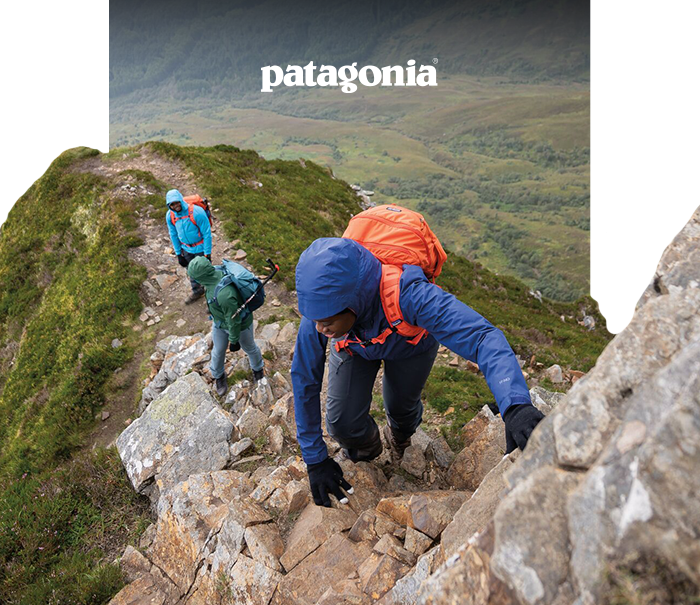 Three hikers in colourful Patagonia jackets and backpacks scrambling up a rocky mountain ridge with a green valley below