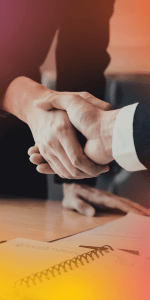 Two professionals shaking hands over a desk with a pen and spiral notebook, sealing a business agreement