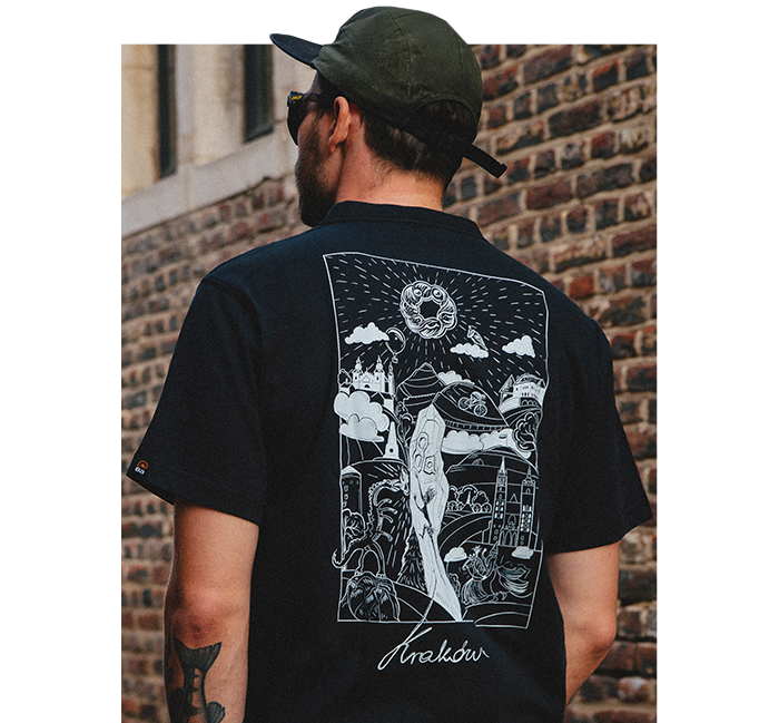 Man wearing black T-shirt with white illustrated Kraków cityscape and central figure on the back, green cap and sunglasses, standing by a brick wall