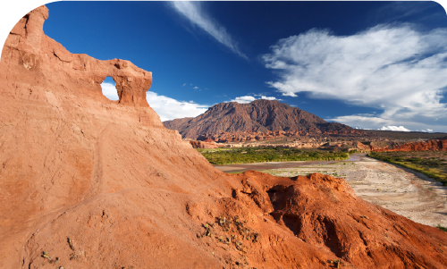 Red sandstone arch and rugged red rock formations overlooking a dry riverbed and distant mountain under a vivid blue sky