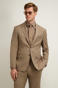 Man in tan suit, matching shirt and tie and sunglasses, standing with one hand in pocket against plain light background