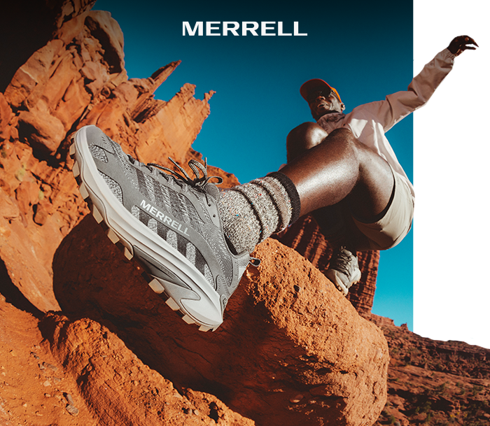 Merrell grey hiking shoe on a hiker's foot leaping over red sandstone rocks in bright desert landscape, brand logo visible