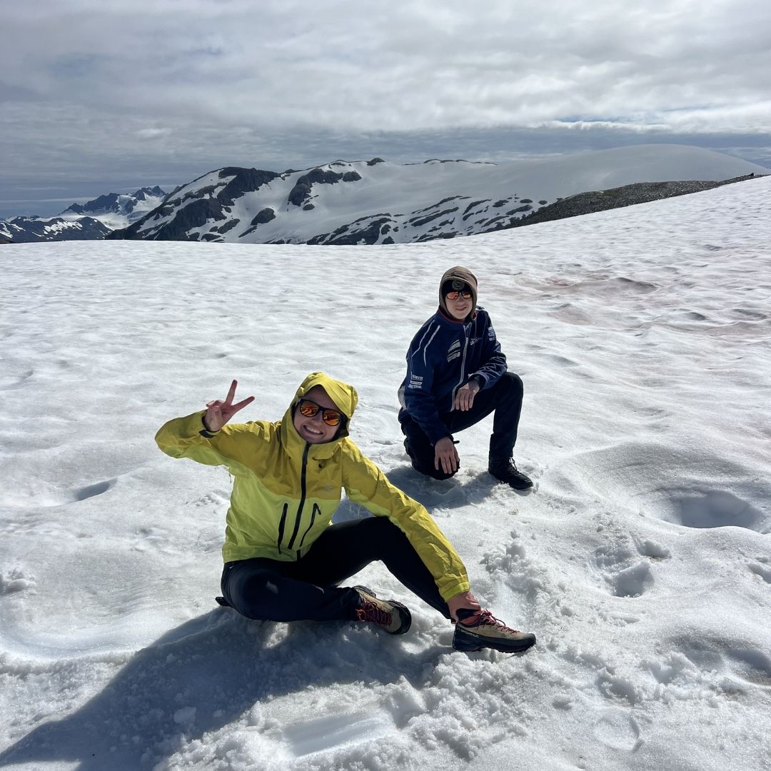 Two hikers in sunglasses and winter jackets pose on a snowy glacier with snow-capped mountains under a cloudy sky
