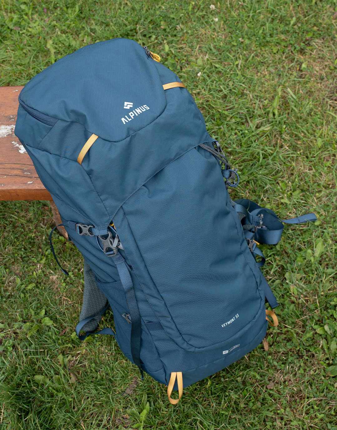 Blue Alpinus Vermont 55 hiking backpack with straps and top lid leaning against a wooden bench on green grass