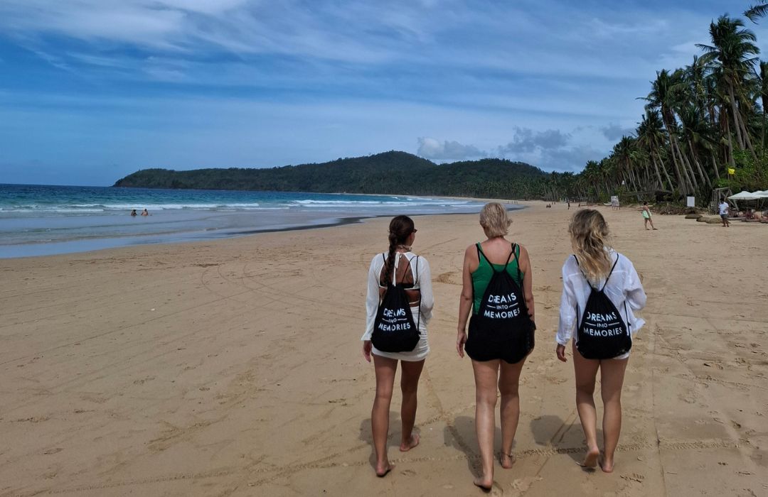 Three women walking along a tropical palm-lined beach, barefoot with matching drawstring backpacks, ocean and distant headland ahead