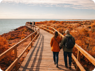 Couple walking hand-in-hand on wooden coastal boardwalk amid autumn foliage, sea and horizon under a pale blue sky