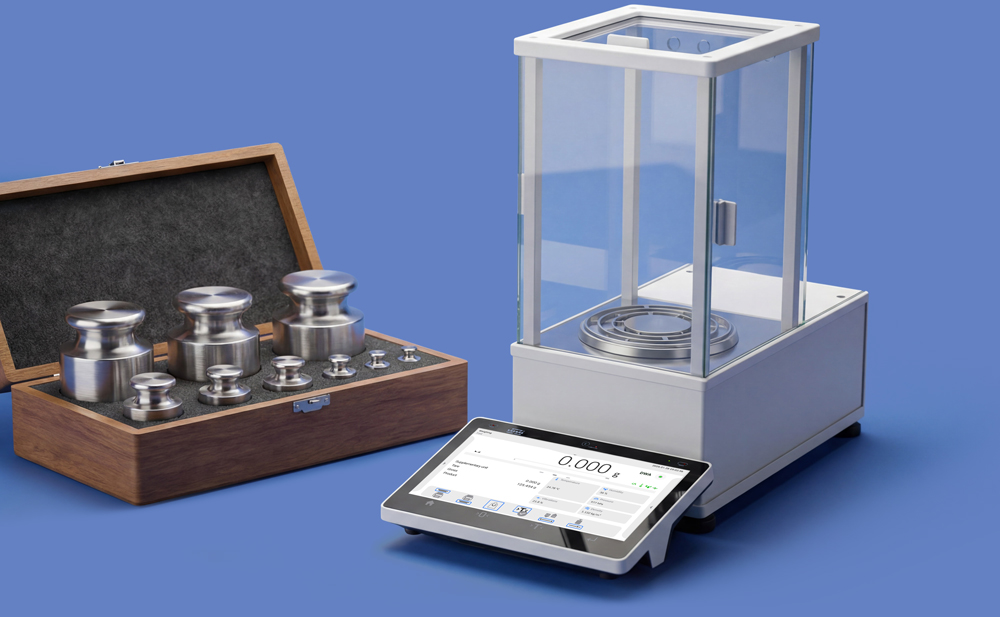 Analytical laboratory balance with glass draft shield and touchscreen display beside a wooden case of stainless steel calibration weights