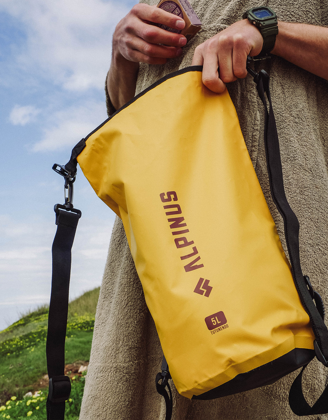 Yellow 5L Alpinus dry bag with shoulder strap held by a person outdoors near grassy hillside and cloudy sky