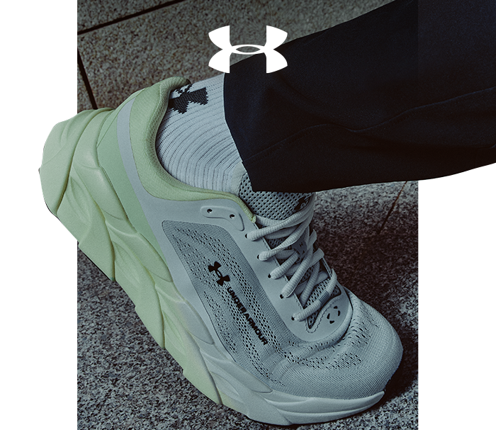 Under Armour pale green and grey running shoe on foot, cushioned sole and mesh upper, worn with logo socks on stone pavement