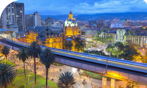 Evening Medellín cityscape showing illuminated Palace of Culture, elevated metro line and lively park with people