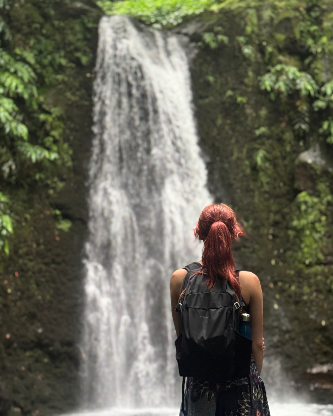 Woman with red hair and backpack seen from behind, gazing at a tall tropical waterfall surrounded by lush green foliage
