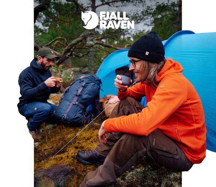 Two hikers in outdoor gear sitting by a blue tent in a pine forest, drinking hot drinks beside backpacks — Fjällräven logo visible