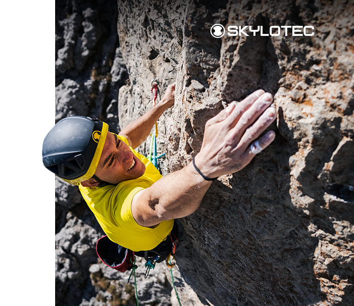 Top-down view of a climber in a yellow shirt and helmet gripping a steep limestone rock face, Skylotec logo visible