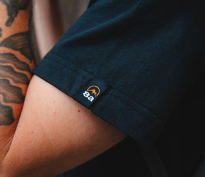 Close-up of tattooed forearm wearing a black t-shirt sleeve with a small tag showing an orange mountain logo and '8a' branding