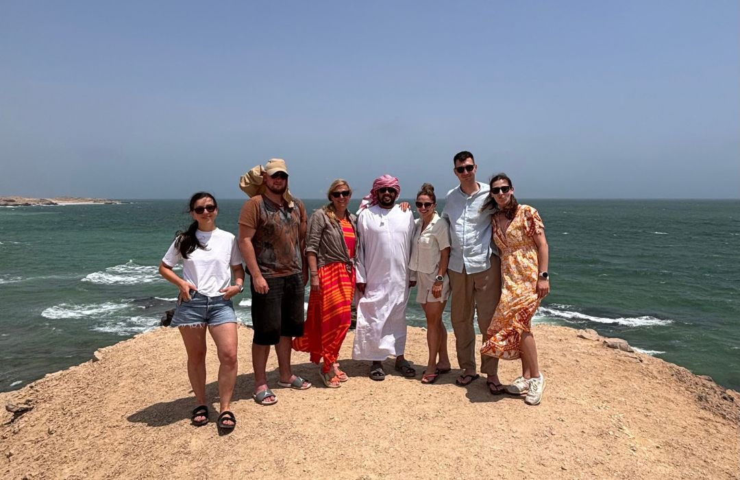 Group of seven tourists, including a man in a white thobe and red headscarf, standing on a sandy cliff overlooking a turquoise sea under a clear sky