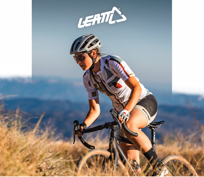 Cyclist wearing helmet, sunglasses and cycling kit riding a gravel bike on a grassy trail with distant hills and a Leatt logo above