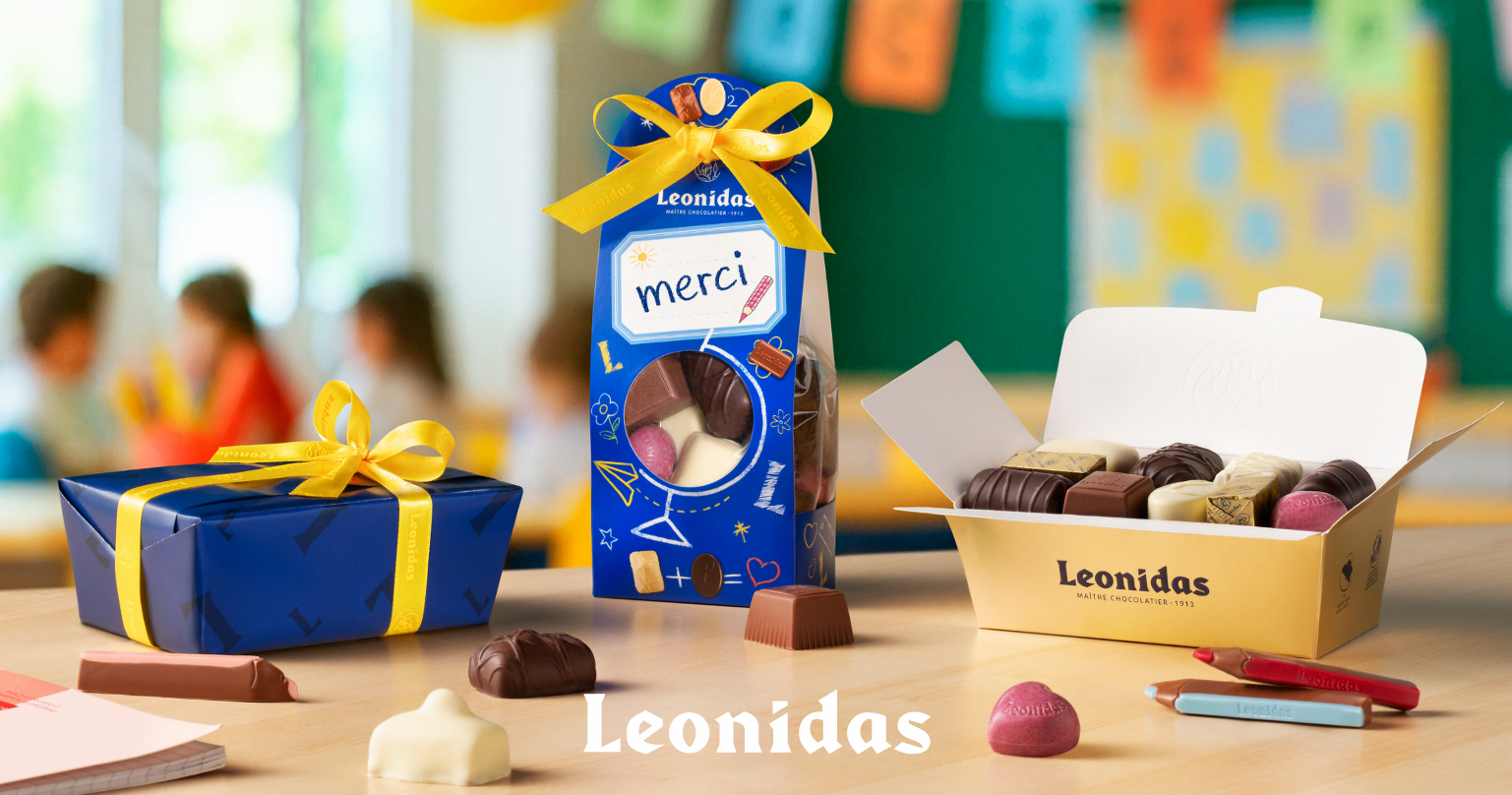 Leonidas gift-wrapped blue box, blue merci bag and open gold carton of assorted chocolates and pralines on a table, blurred classroom background