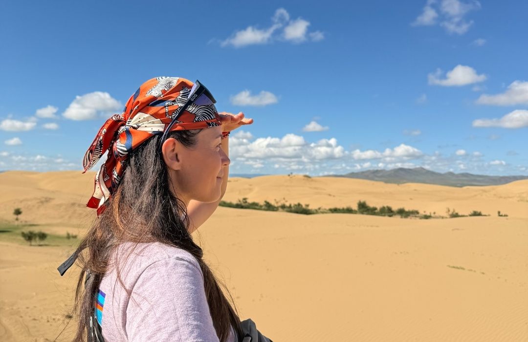 Young woman wearing a patterned headscarf and sunglasses, shielding her eyes as she looks across golden sand dunes under a bright blue sky