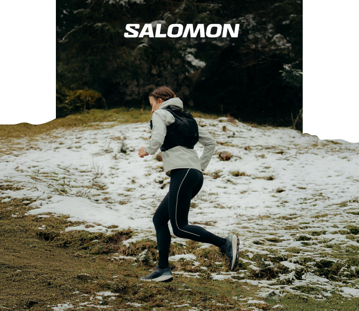 Person trail running on a snowy grassy hill wearing Salomon jacket, backpack and trail shoes beneath a forest with SALOMON logo above