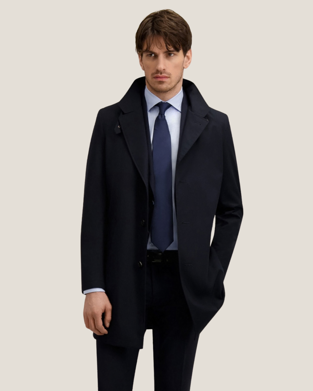Male model wearing a navy single-breasted overcoat with light blue shirt and navy tie on neutral background