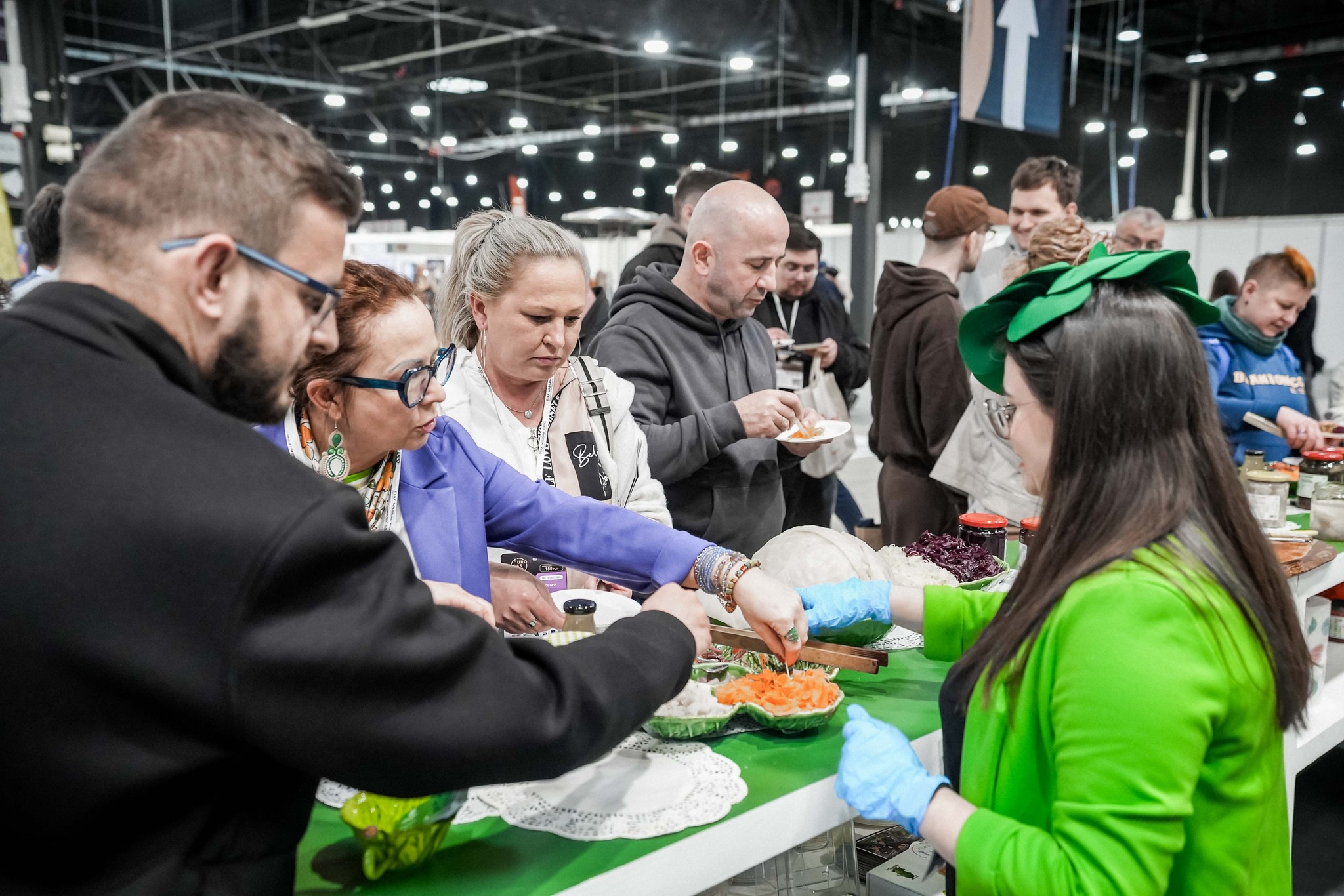 People sampling pickled vegetables at a bustling indoor food market stall, server in green jacket and gloves offering samples