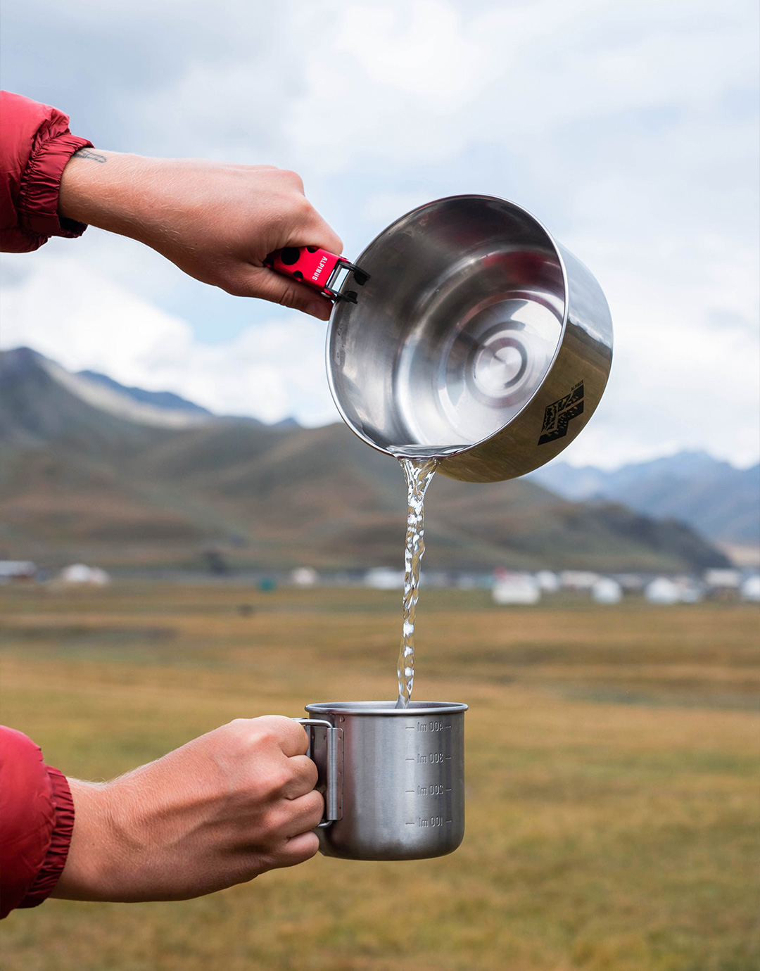 Hands pouring water from a stainless-steel camping pot into a metal mug at a mountain campsite under a cloudy sky